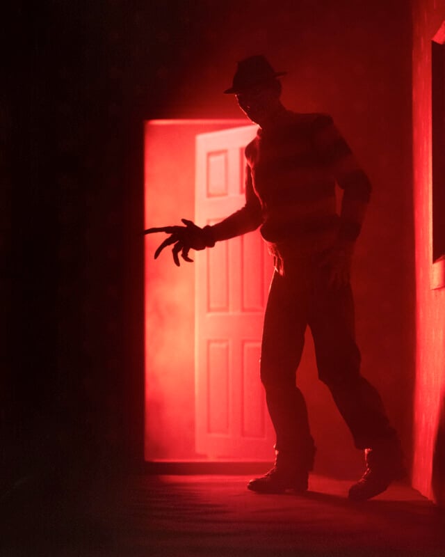 A shadowy figure with a glove of long sharp blades stands menacingly in front of an open door glowing with eerie red light, creating a suspenseful and ominous atmosphere.