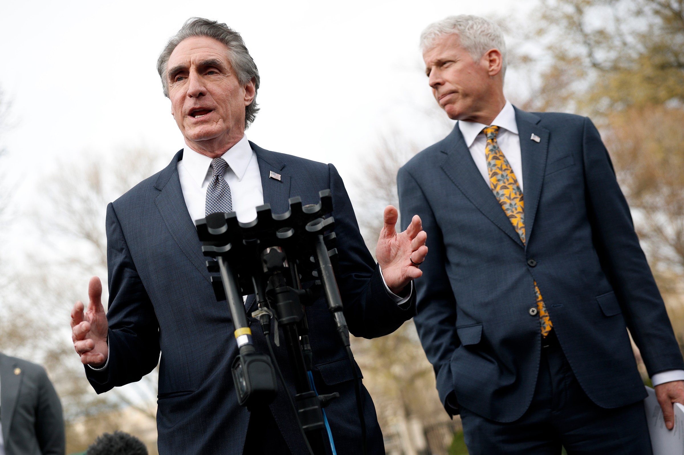 Seen from a low angle below, Doug Burgum, in a navy suit, talks into a gaggle of press microphones, with another man in a navy suit standing next to him.