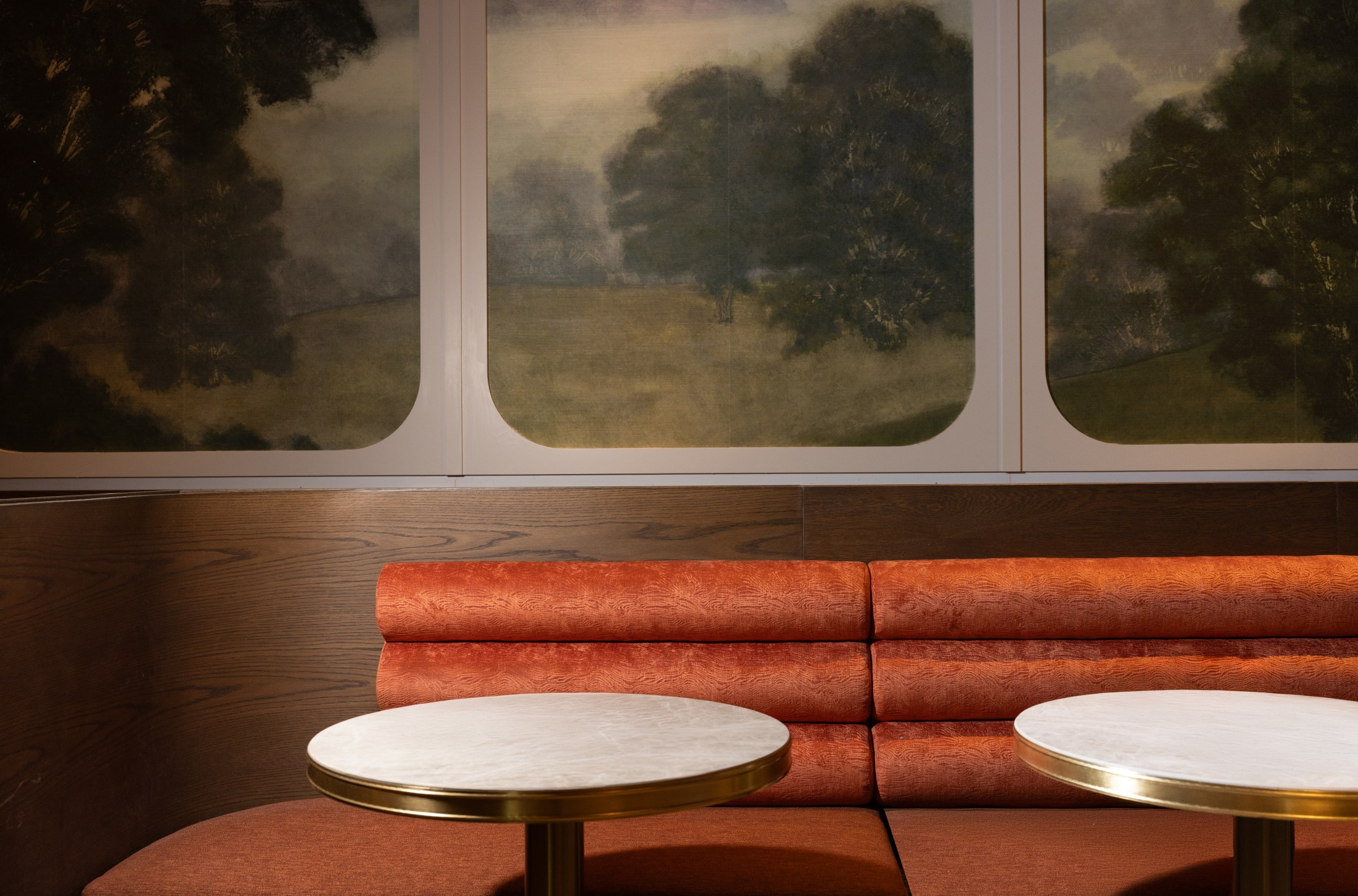 A restaurant dining room with an orange banquette against the wall, white round tables, and a countryside landscape painting on the wall.