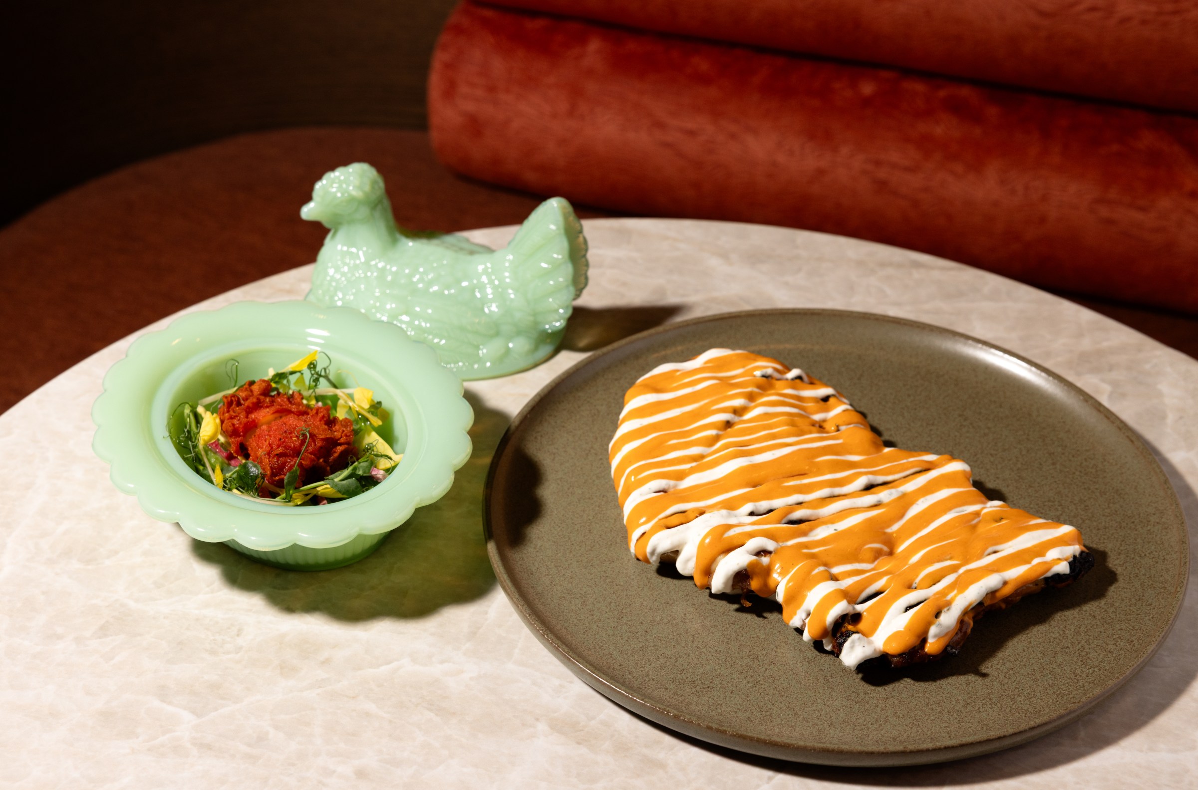 A green bowl with a crispy chicken and greens next to the chicken-shaped cover next to a plate of chicken with sauce.