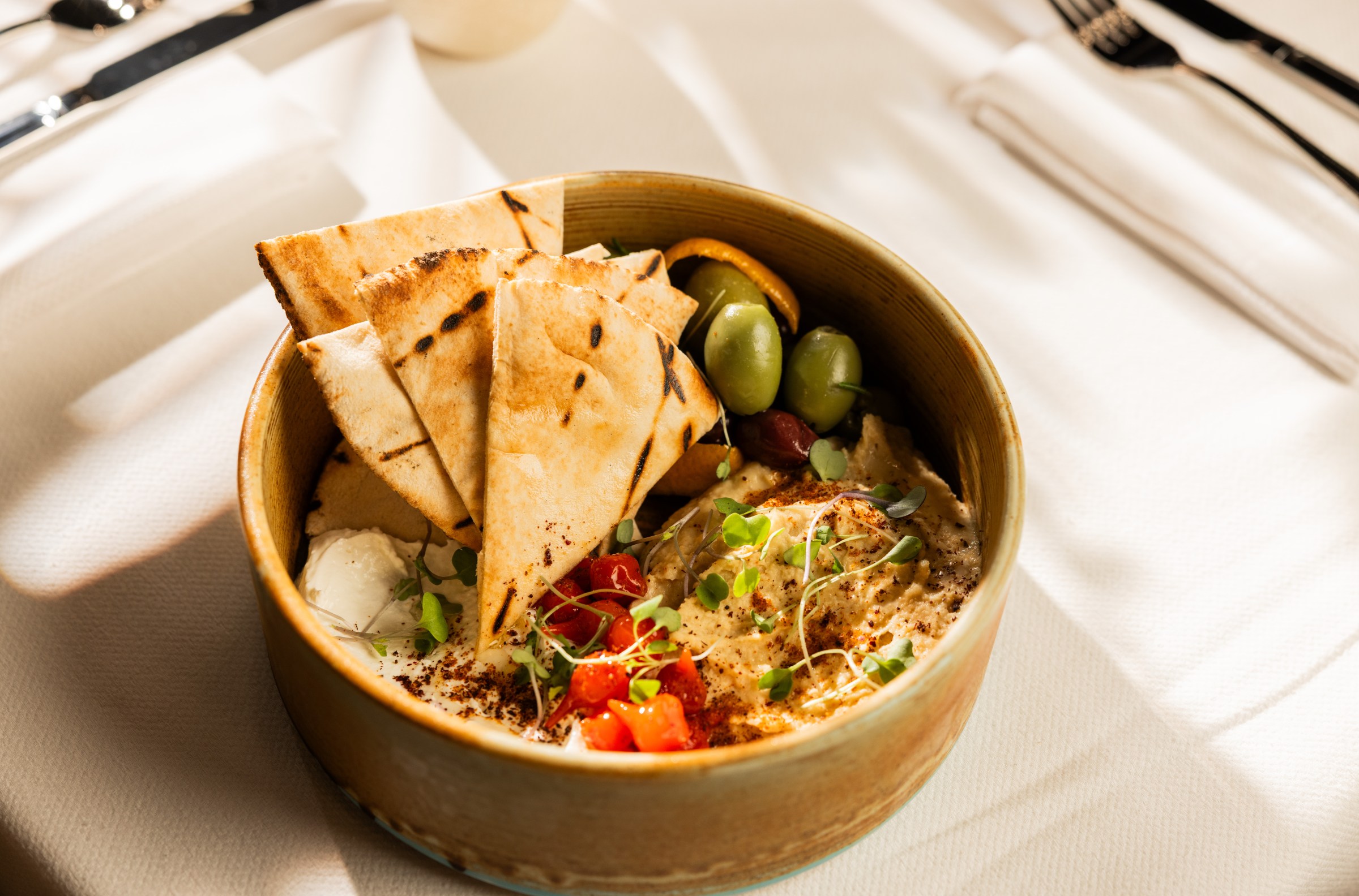 A bamboo bowl holds hummus, olives, vegetables, and pita bread.