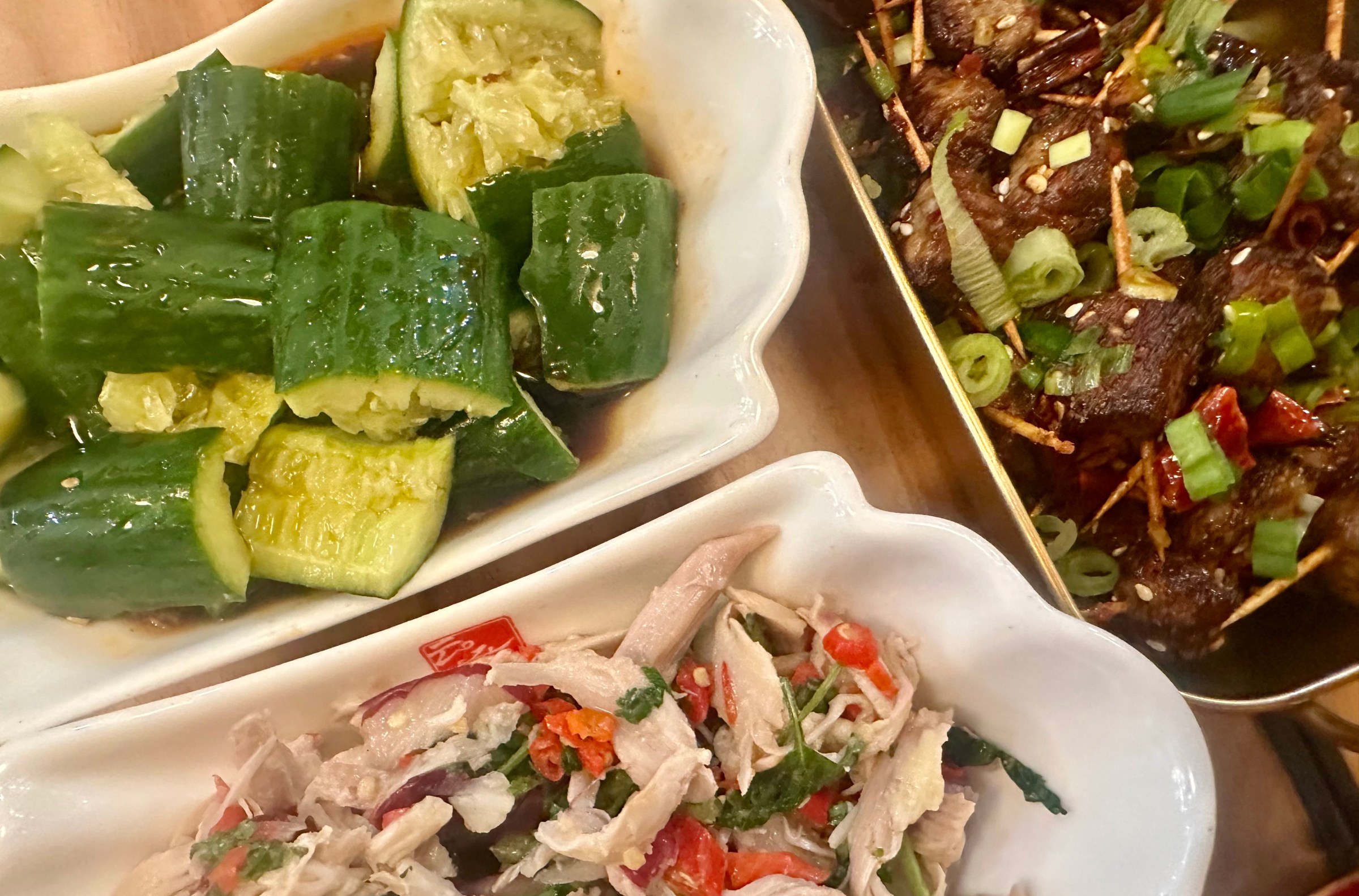 A dish of chunky sliced cucumbers, another with diced beef and green onions, and a third with shredded chicken and vegetables.