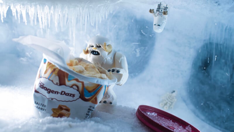 A LEGO yeti figure grabs ice cream from an open Häagen-Dazs tub in an icy cave, with another small LEGO creature in the background and a red lid on the snow.