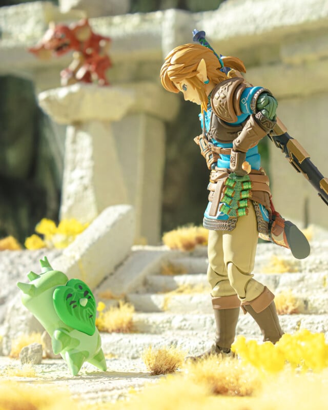 A toy figure of Link from The Legend of Zelda stands on stone steps outdoors, looking down and smiling at a small, green, leafy Korok figurine. Bright sunlight and yellow grass surround the scene.