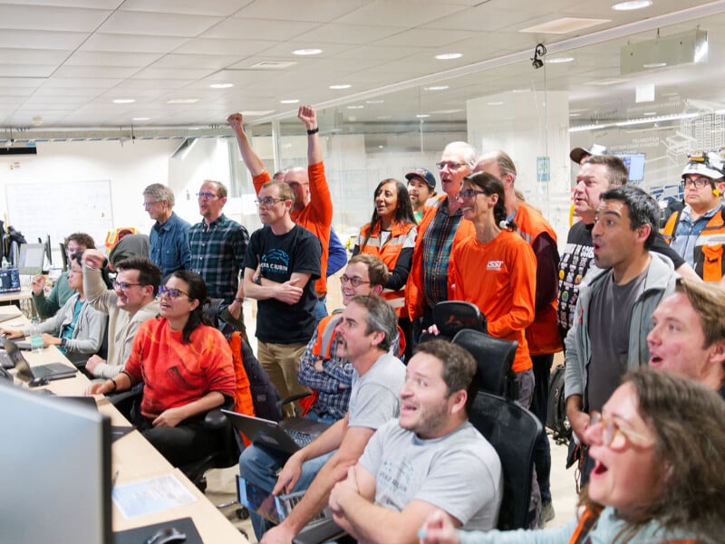 A group of excited people in an office environment watch computer screens, some cheering and smiling, while others react with surprise and joy. The scene appears to capture a celebratory or victorious moment.