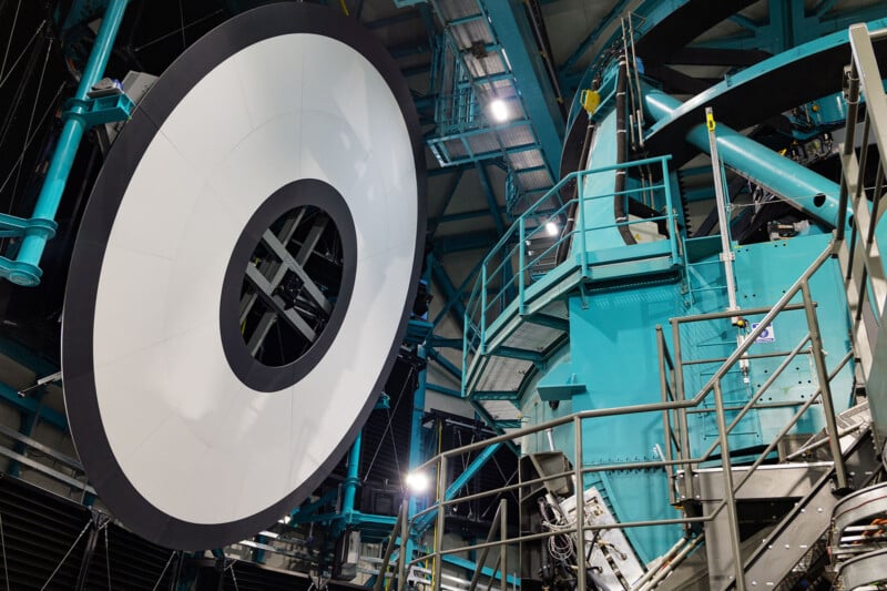 A large, circular telescope mirror with a central hole is mounted inside an industrial facility, surrounded by teal metal beams, platforms, and railings.
