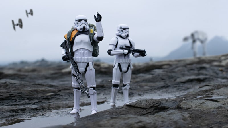 Two stormtroopers stand on rocky terrain under a cloudy sky. One raises a hand while the other holds a blaster. TIE fighters fly in the background, and an AT-AT walker is visible in the distance.