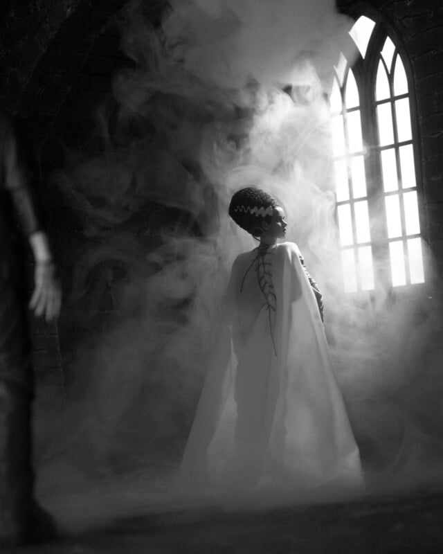 A woman in a dramatic white cloak stands in swirling fog, facing a tall arched window with light streaming in. The scene is dark and mysterious, with gothic architectural details in the background.