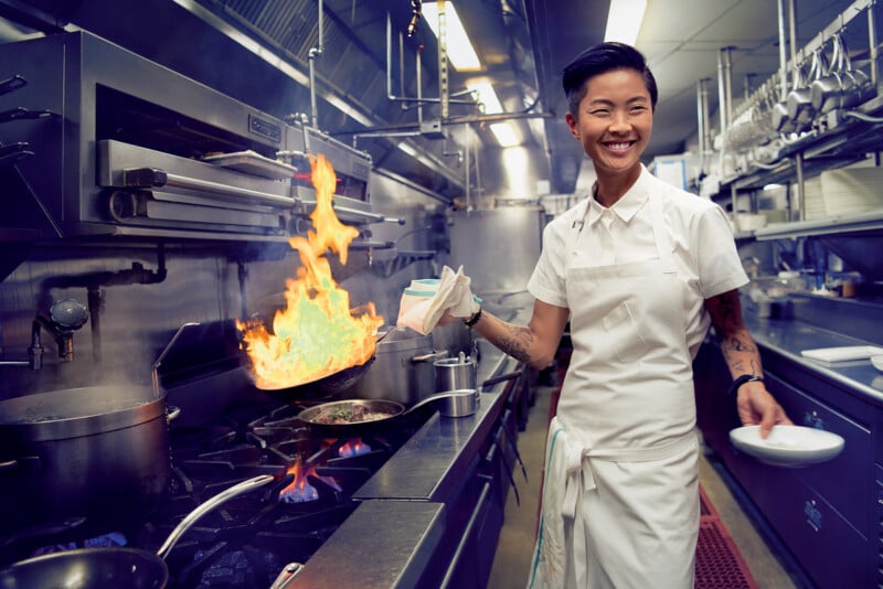 A chef in a white uniform smiles while cooking in a commercial kitchen, holding a towel and plate. Flames rise from a pan on the stove, and pots and utensils are visible around the kitchen.