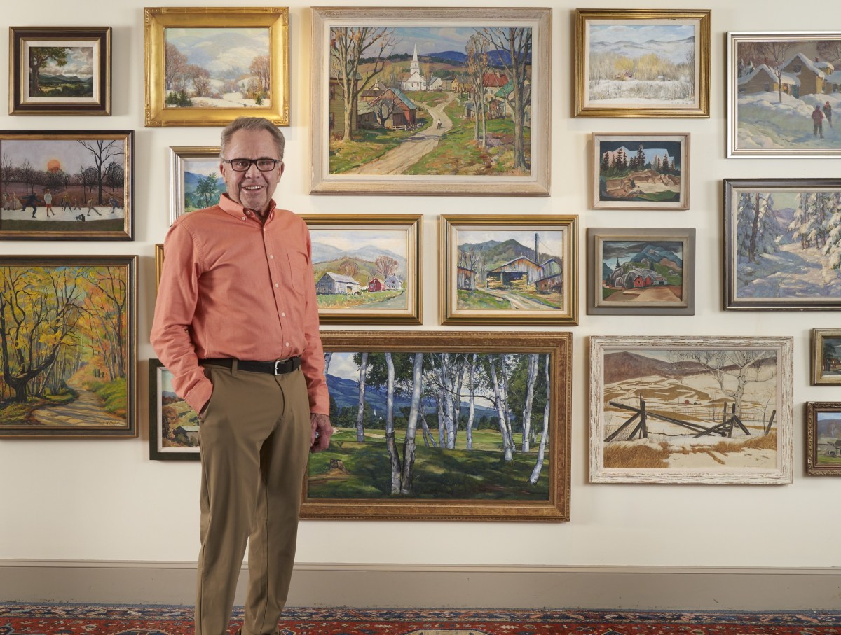 A man in an orange shirt and khaki pants stands in front of a wall displaying multiple framed landscape paintings.