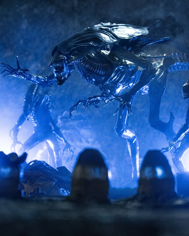 A menacing, biomechanical alien creature with sharp teeth and claws looms in a dark, blue-lit cave, surrounded by similar creatures. Two blurry helmeted figures are visible in the foreground.