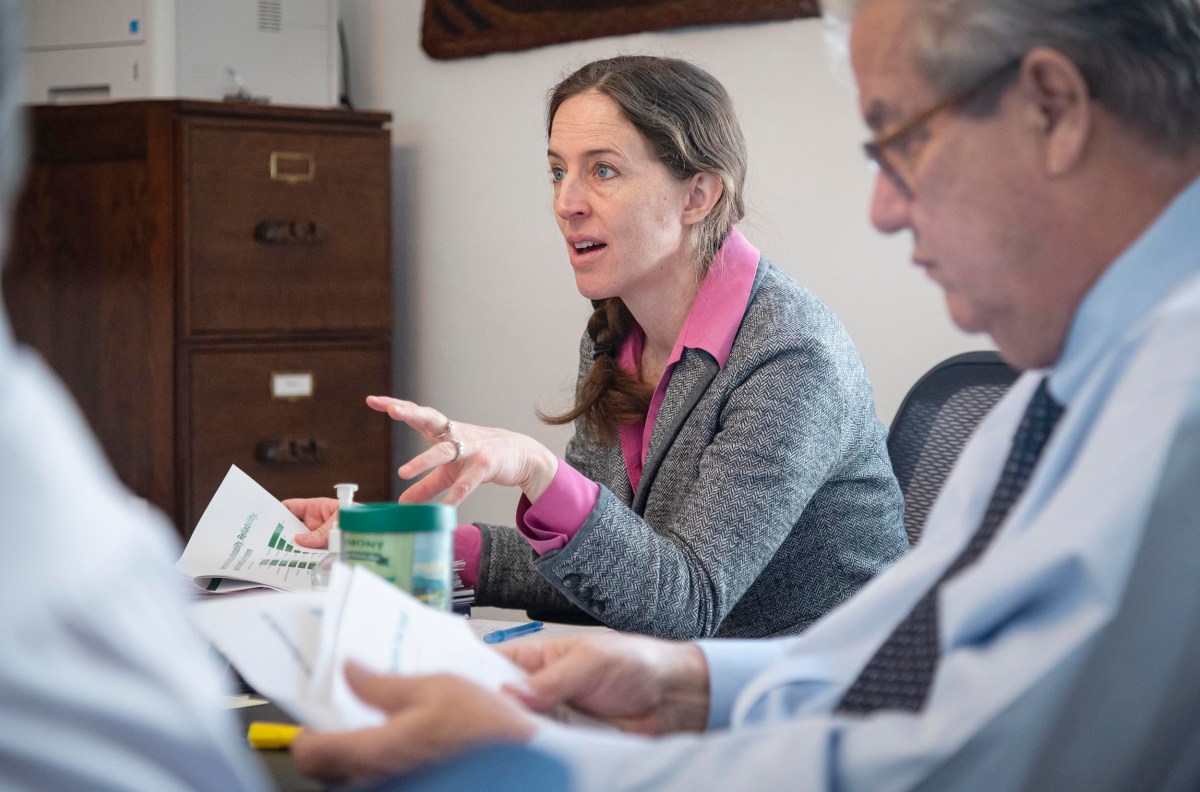A woman in a gray blazer talks during a meeting, gesturing with her hand. Two men, one blurred in the foreground and the other beside her, are seated, holding papers. A filing cabinet is in the background.