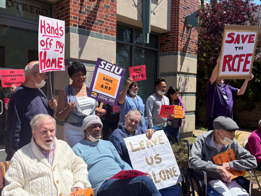A group of people, some seated and some standing, hold signs reading “Hands off my Home,” “Save the RCFE,” and “Leave Us Alone” outside a building during a protest.