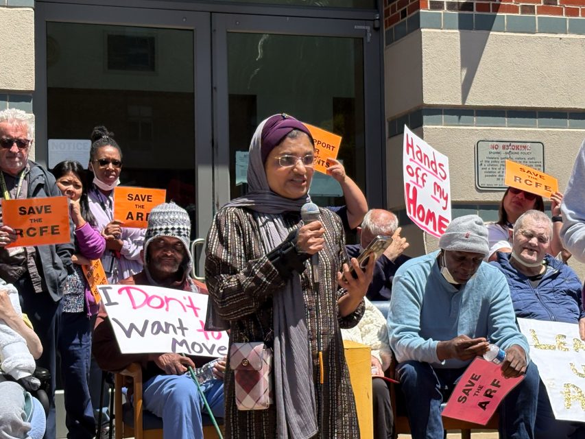 A woman speaks into a microphone at a protest outside a building, surrounded by people holding signs that read “SAVE THE RCFE” and “Hands off my Home.”.