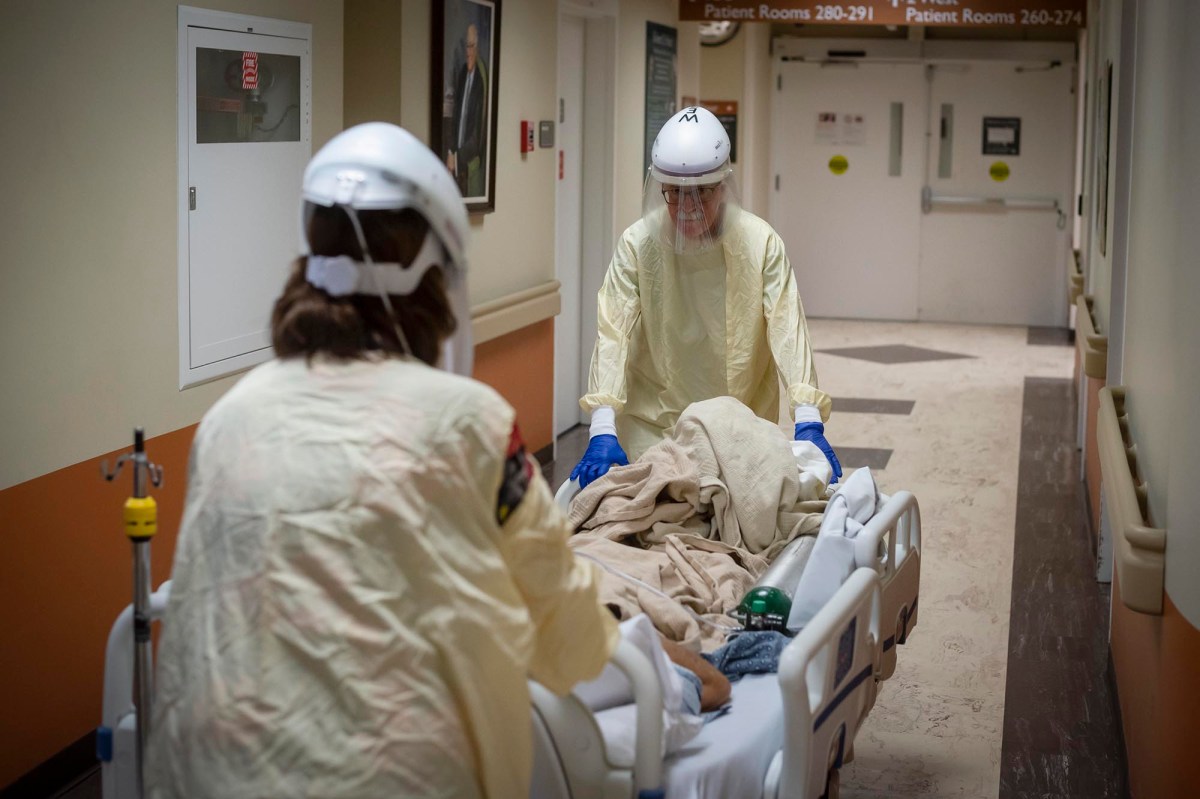 Two medical staff in protective gear push a patient on a hospital bed down a hallway. The patient is covered with a blanket. The hallway leads to a series of patient rooms.