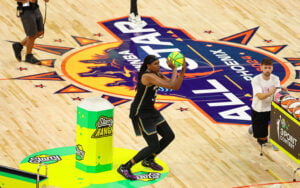 Basketball player shooting during an WNBA 3-point contest, standing near a "Starry Range" stand on the court.