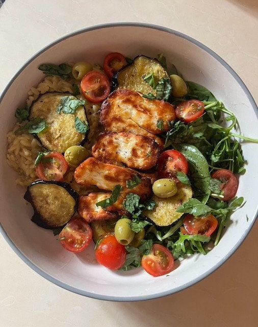 Bowl of orzo pasta with grilled halloumi, roasted eggplant, cherry tomatoes, olives, and greens.