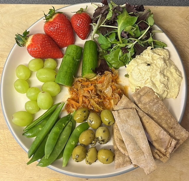 Plate of food with hummus, pita bread, kimchi, olives, grapes, strawberries, cucumbers, and sugar snap peas.