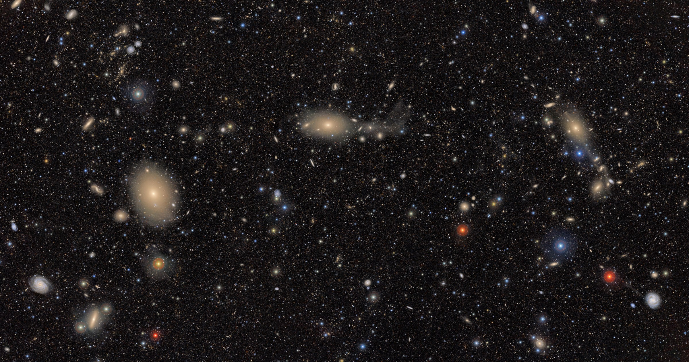 A wide-field image of deep space showcasing the Virgo Cluster, teeming with galaxies of various shapes, sizes, and colors. Large elliptical galaxies appear as soft yellowish glows, while spiral galaxies display distinct arms. Numerous smaller galaxies, star-like points, and reddish and bluish dots are scattered throughout the black cosmic background. A faint tidal stream trails off one of the galaxies near the center-right, suggesting gravitational interaction. The scene is a rich, visually dense tapestry of galactic evolution and structure.