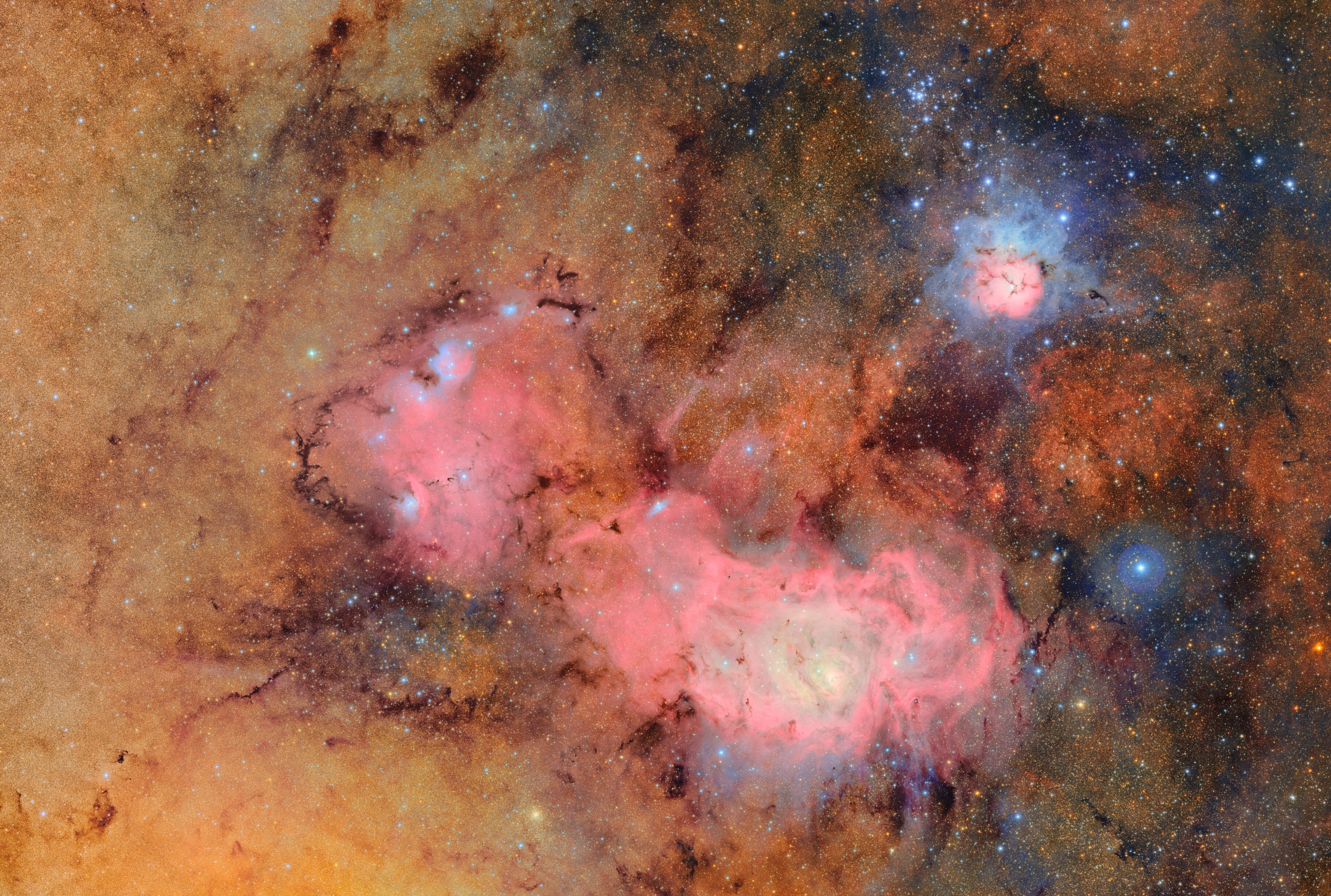 A vivid, high-resolution image of a star-forming region in deep space, prominently featuring the Lagoon Nebula (Messier 8) in glowing pink and the Trifid Nebula (Messier 20) in a blend of pink and blue hues. The nebulae are surrounded by dense star fields and golden interstellar dust clouds. Dark filaments of cosmic dust snake through the frame, especially near the pink emission areas. The overall color palette includes warm golds, reds, cool blues, and purples, highlighting active regions of stellar birth and ionized gas.