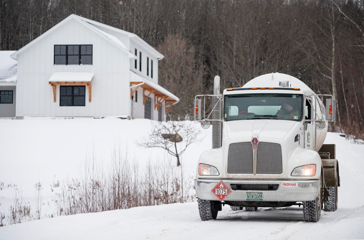 A propane truck drives on a snowy road toward a white house with wooden accents surrounded by trees.
