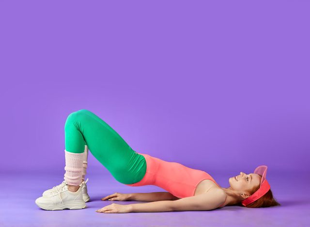 ginger girl making hip bridge exercise, also known glute bridge to work out glutes and abs muscles, lying on floor, both legs knees bent, feet and hands on ground, dressed colorful aerobics clothes