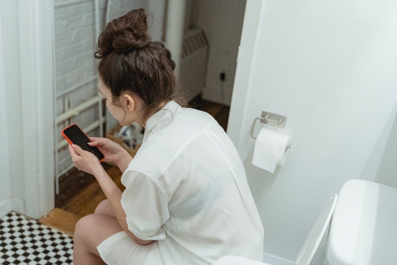 Woman wearing white shirt using phone while on toilet seat