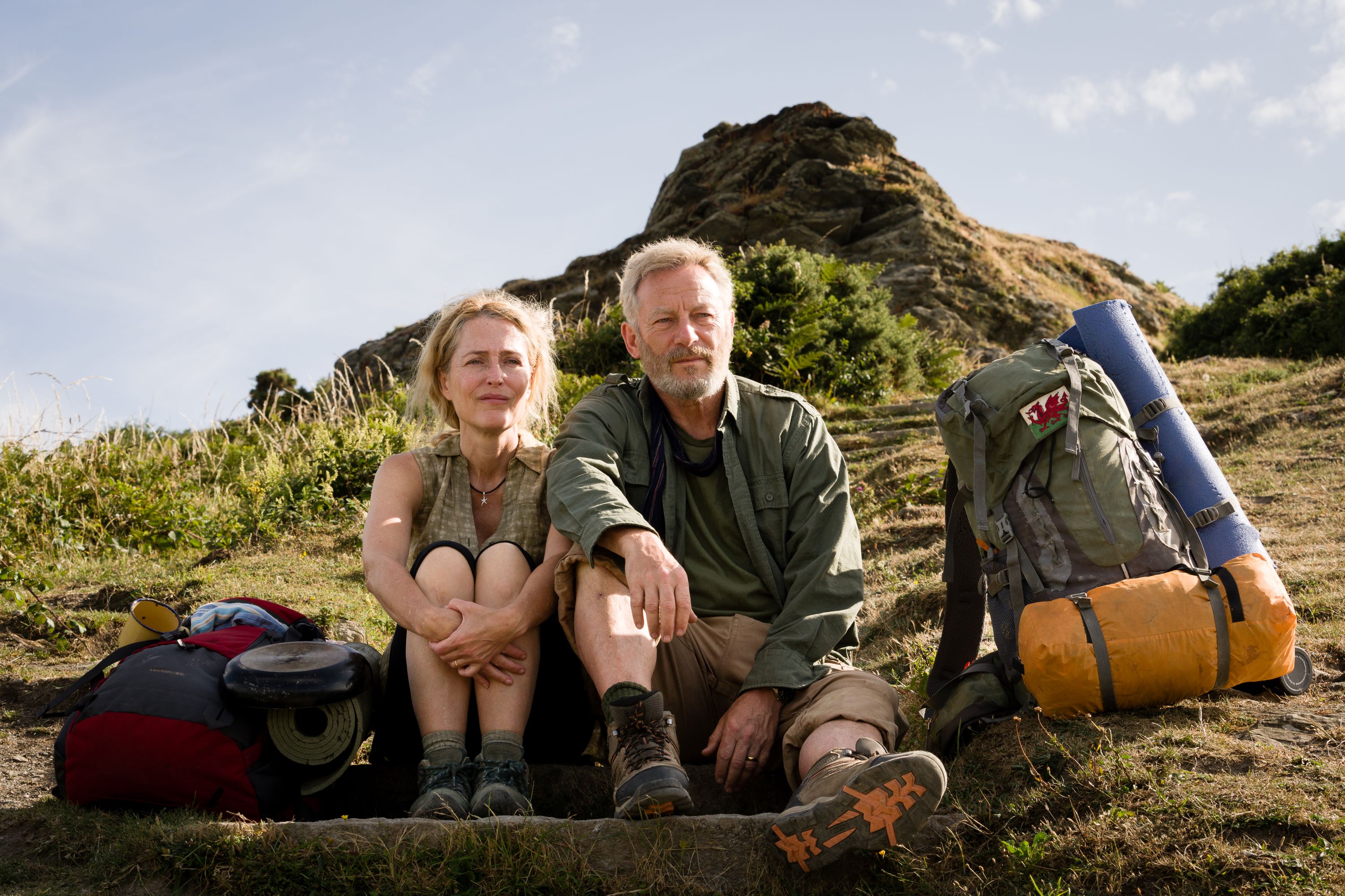 Film still of a couple sitting on a hillside with backpacks.