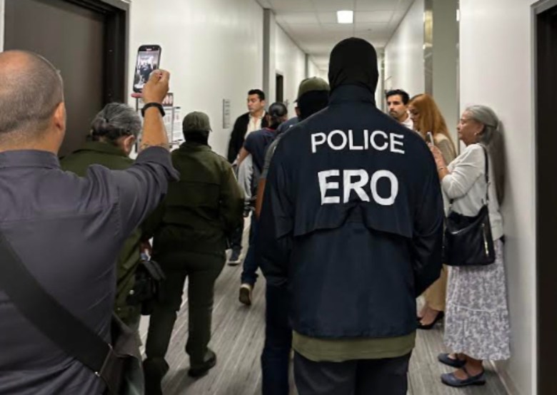 Several people crowd a hallway, some with cellphones in hand, while an ICE enforcement officer stands sentry.