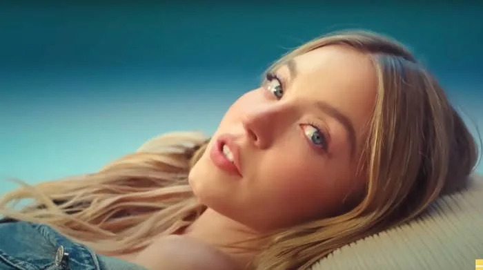 Person reclining, gazing slightly at the camera with a relaxed expression, wearing an off-shoulder denim outfit. Hairstyle is loose and wavy
