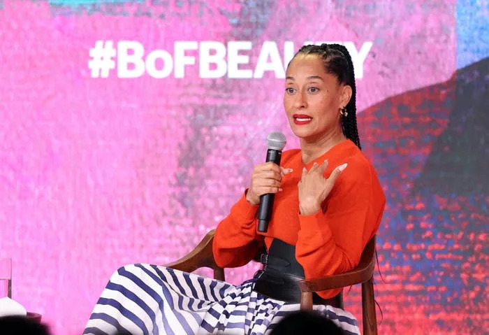Tracee Ellis Ross speaking at an event, wearing a bright top and striped pants, holding a microphone in front of a backdrop with "#BoFBEAUTY" text