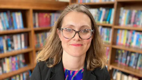 BBC Head teacher Louise Cowley in front of bookshelves in a library. She has long blonde hair and glasses.