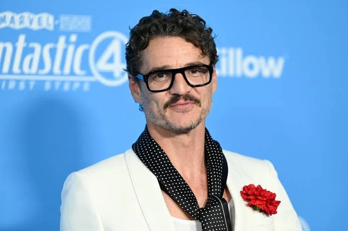 Man in a white suit and polka dot scarf on a blue background, wearing glasses and a red flower on lapel, at a celebrity event