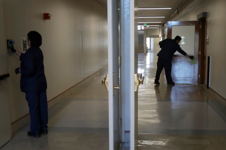 Two janitorial workers using rags to scrub the walls and doors of a school hallway. The center of the photo is divided by two doors while the workers stand on opposite sides of hall scrubbing.
