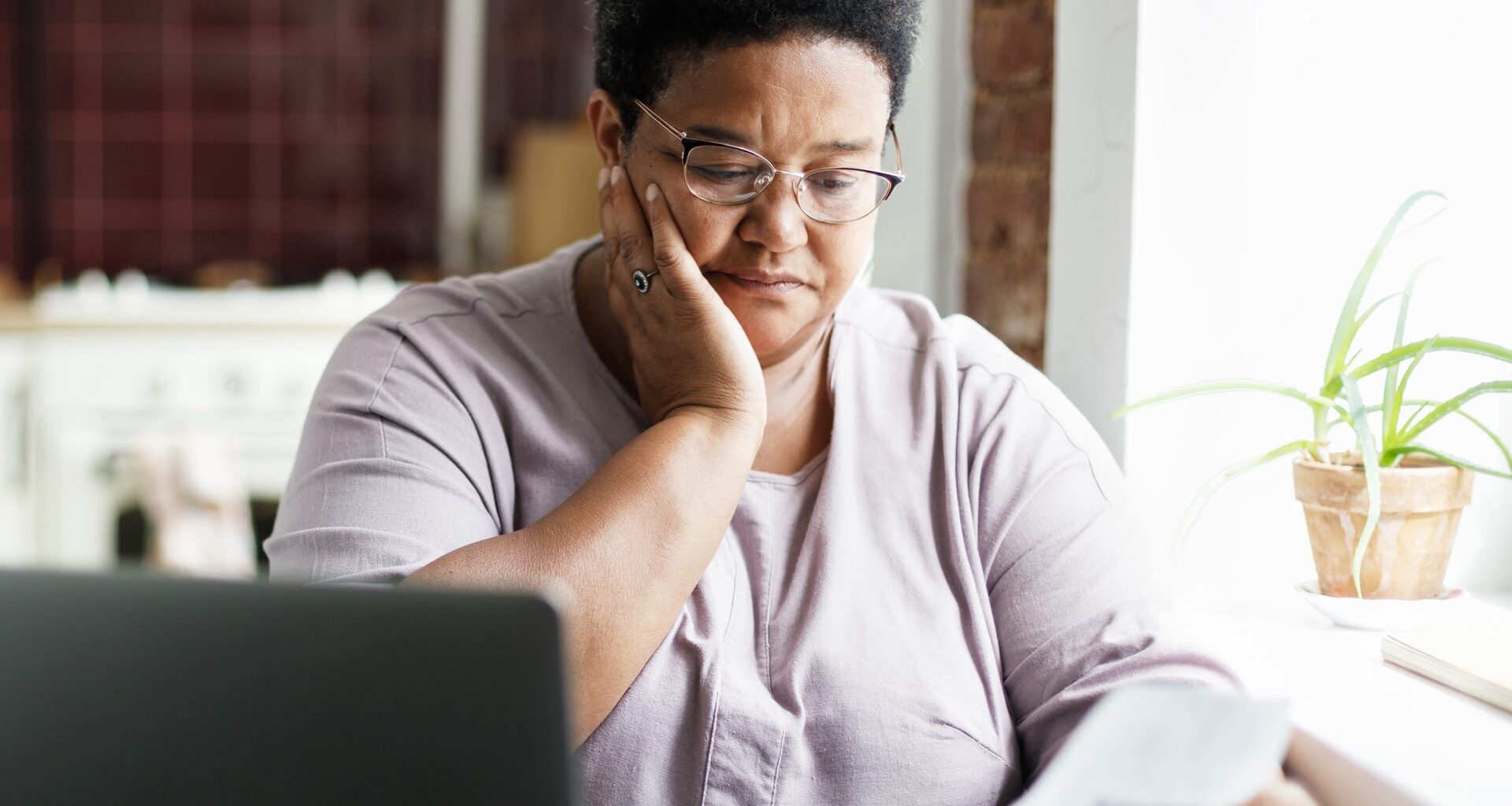 A mature African American woman in glasses touches her cheek, looking sad and puzzled by a high utility bill.