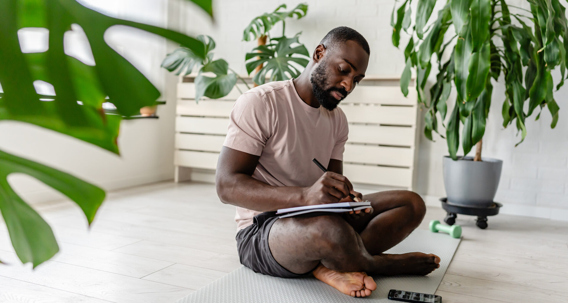 Young African-American man sitting cross-legged on a yoga mat at home, writing in a notebook after a workout, with a smartphone nearby, creating a calm and focused atmosphere
