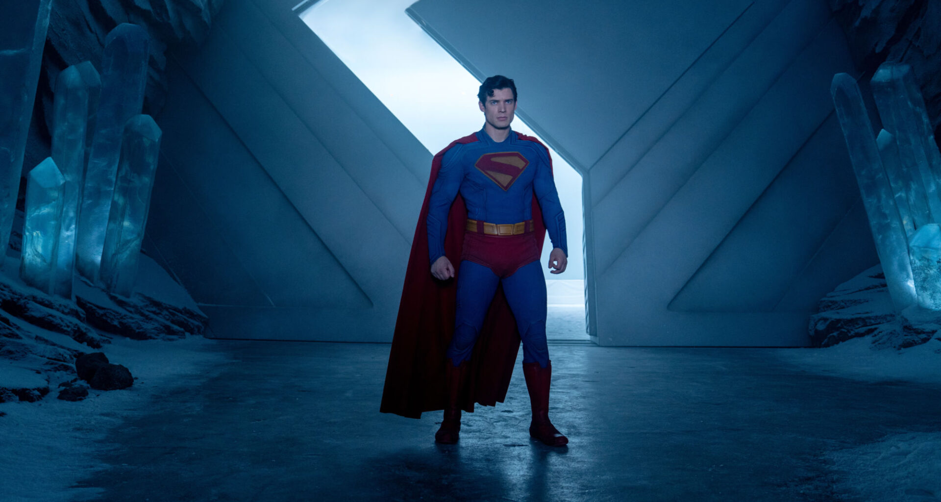 Superman stands confidently in a futuristic ice cave, wearing his iconic blue suit with a red cape. Crystalline structures are visible in the background against a blue-lit atmosphere.