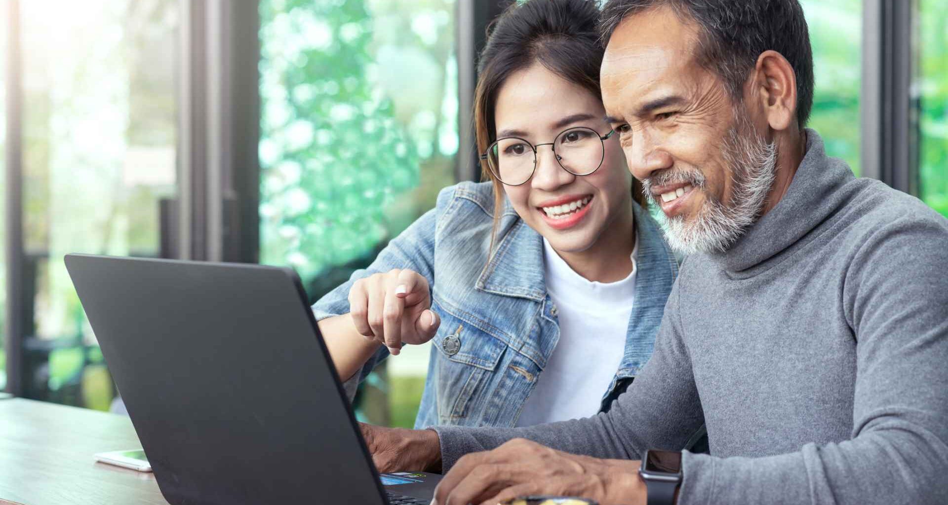Attractive mature asian man with white stylish short beard looking at laptop computer with teenage eye glasses hipster woman in cafe.