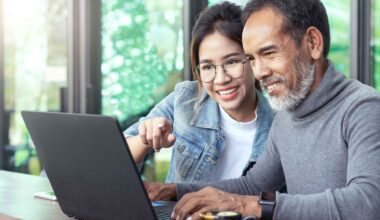Attractive mature asian man with white stylish short beard looking at laptop computer with teenage eye glasses hipster woman in cafe.