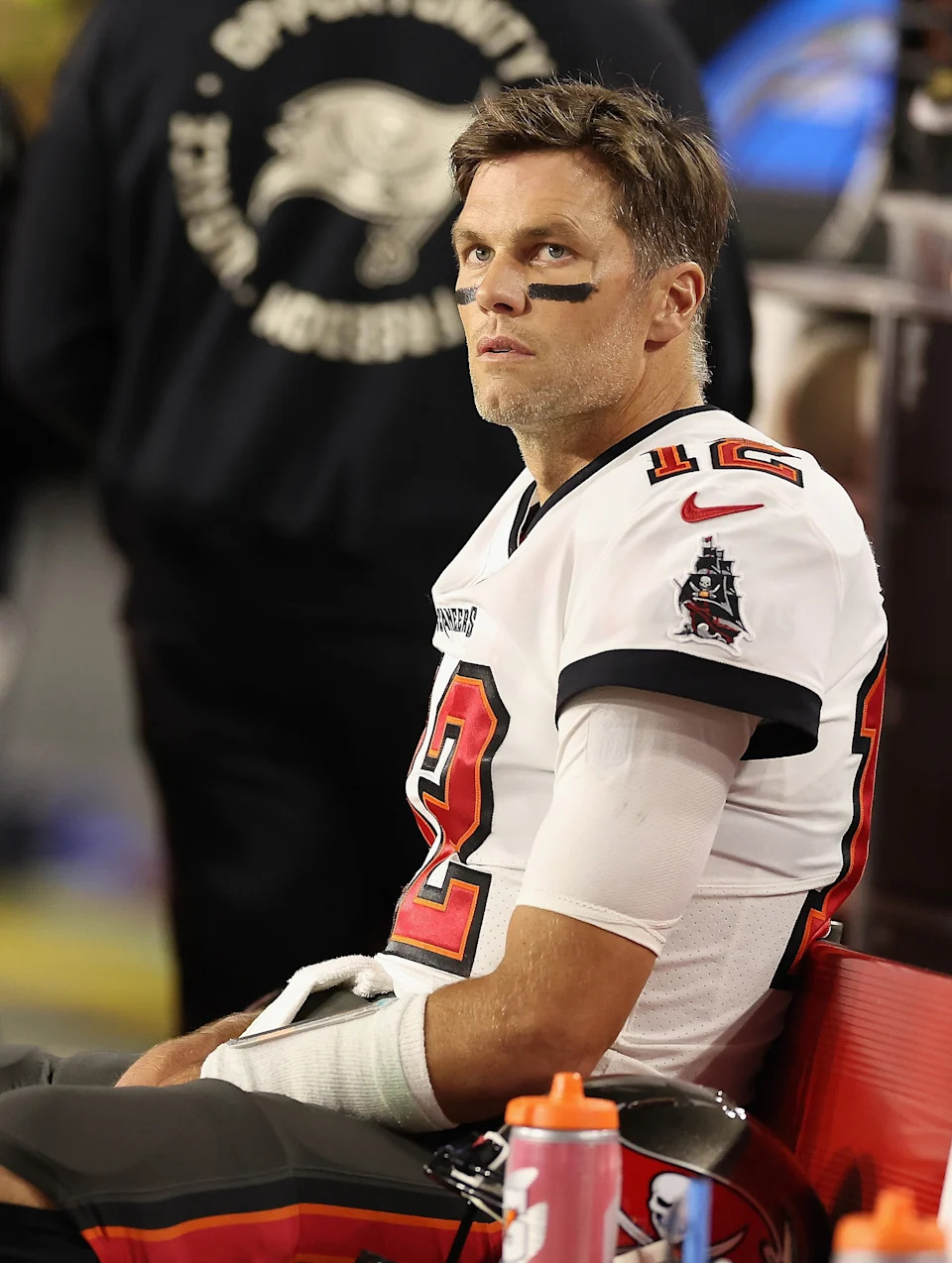 Football player in uniform sitting on the sidelines, looking focused. Number 12 jersey, with eye black on. Water bottles nearby