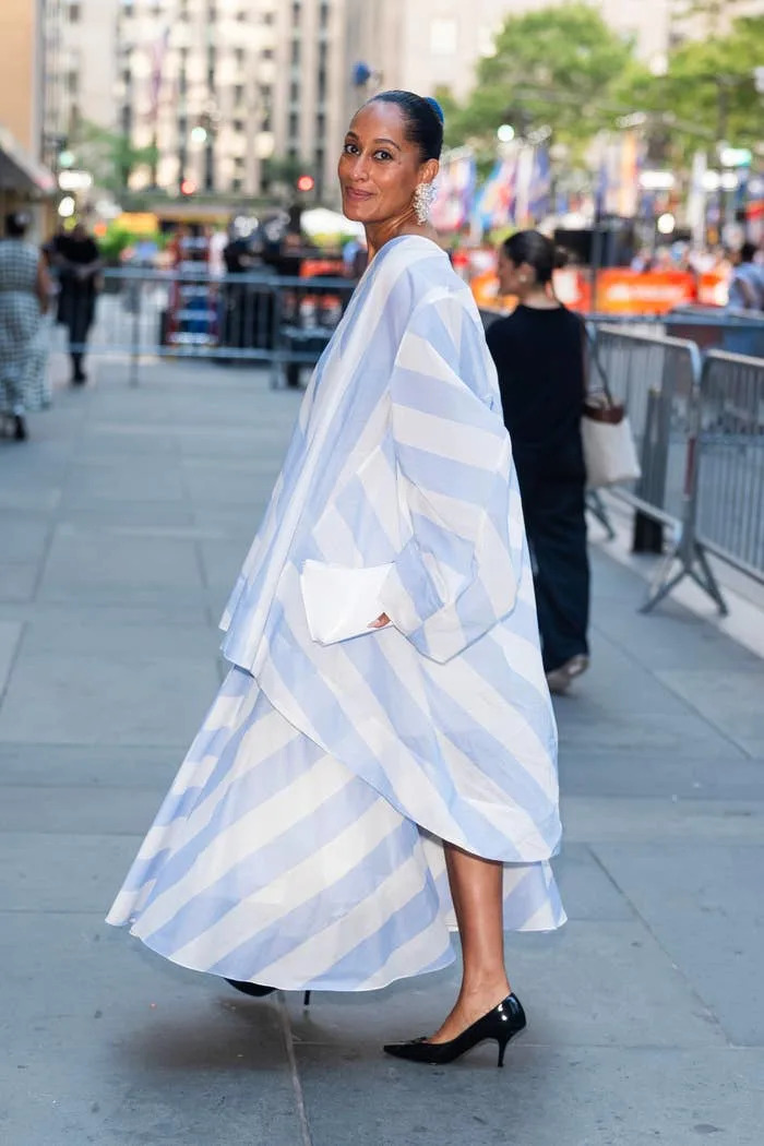 Tracee Ellis Ross wearing a flowing, striped outfit and heels poses outdoors, turning to smile at the camera