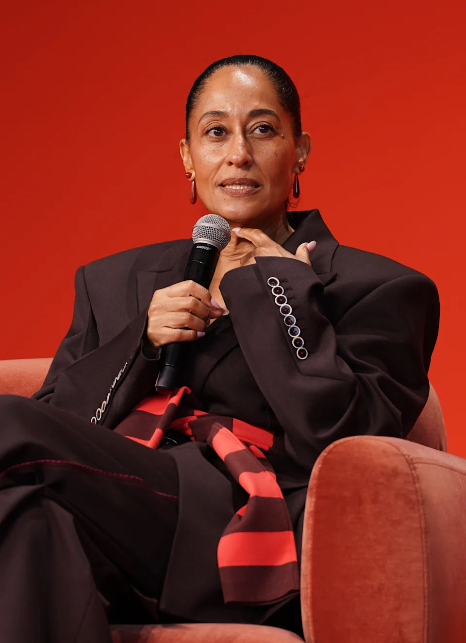 Tracee Ellis Ross seated, wearing an oversized suit with a striped detail, holding a microphone, speaking at an event