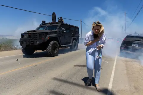 Getty Images A woman in blue jeans with blond hair runs away from a military vehicle and white smoke which looks like tear gas. 
