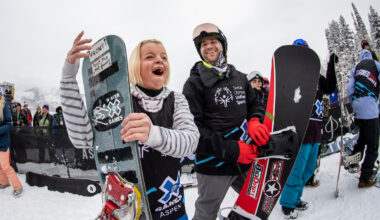 Two snowboarders in winter sports gear smile and pose with their snowboards at a snowy outdoor event with people and trees in the background.