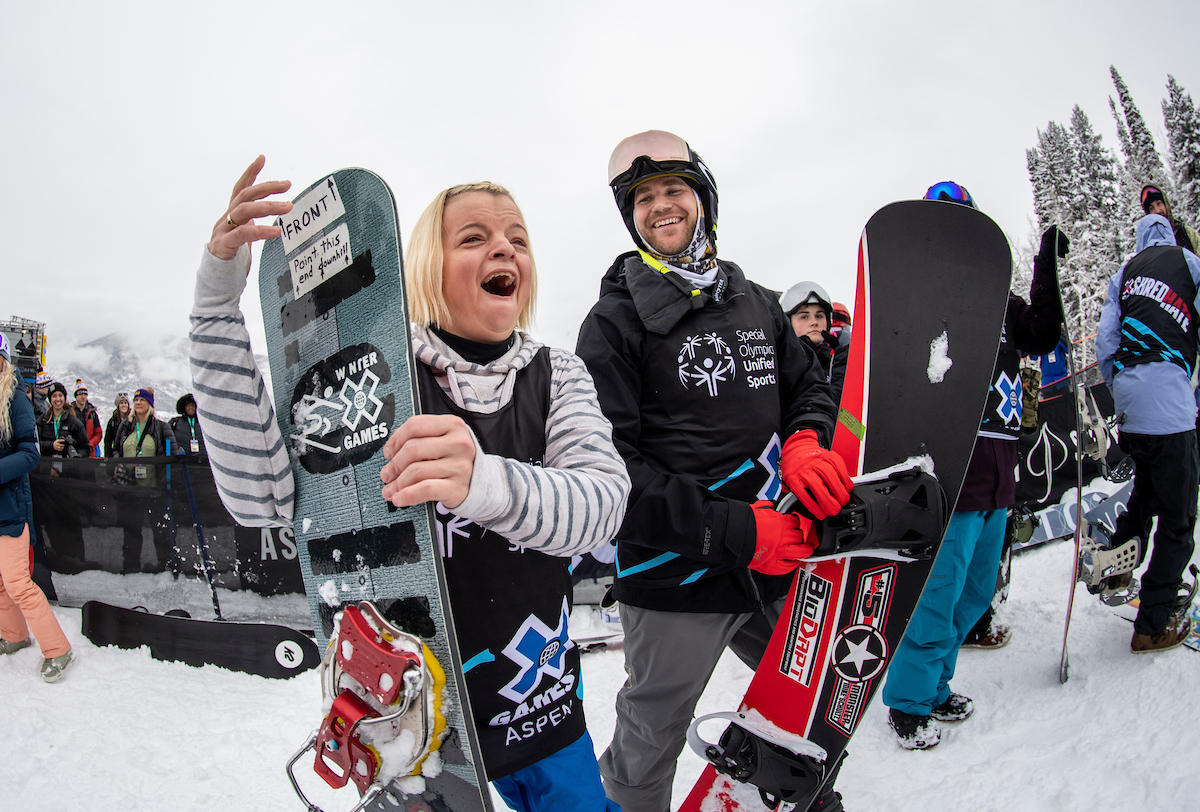 Two snowboarders in winter sports gear smile and pose with their snowboards at a snowy outdoor event with people and trees in the background.