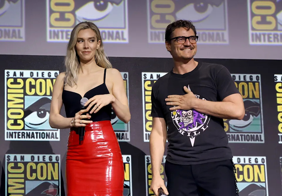 Two actors on stage at Comic-Con. The woman is in a fitted black top and shiny skirt; the man wears a printed T-shirt, smiling with hand on chest