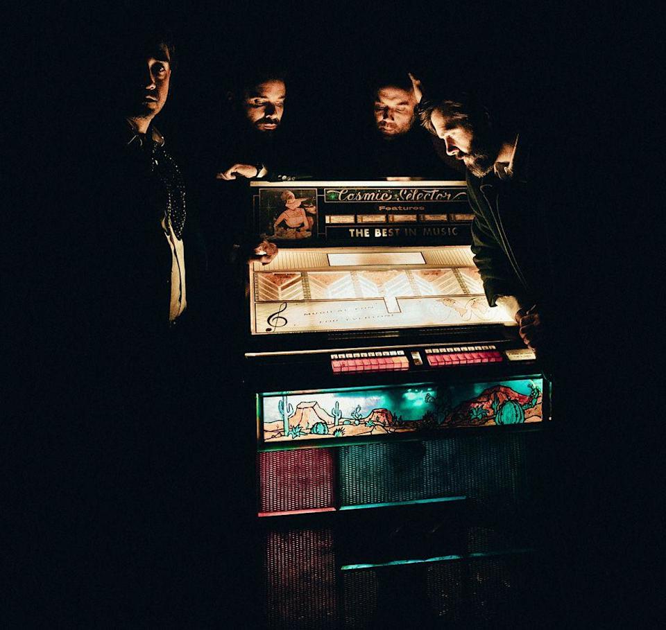 Four band members standing in semi-darkness around an old-fashioned lit-up jukebox with "The Cosmic Selector" written in decorative lettering on the top