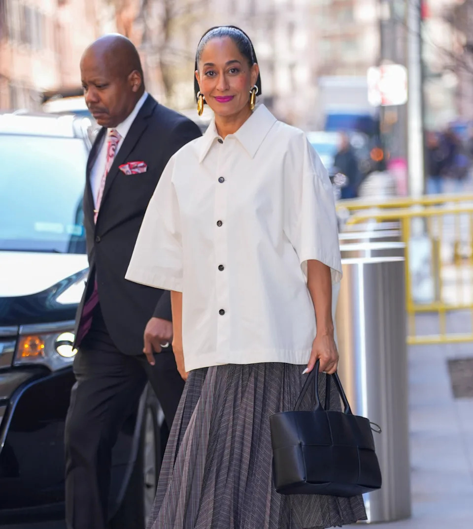 Tracee Ellis Ross in a stylish oversized shirt and pleated skirt walks along a city street, holding a black handbag, accompanied by a man in a suit