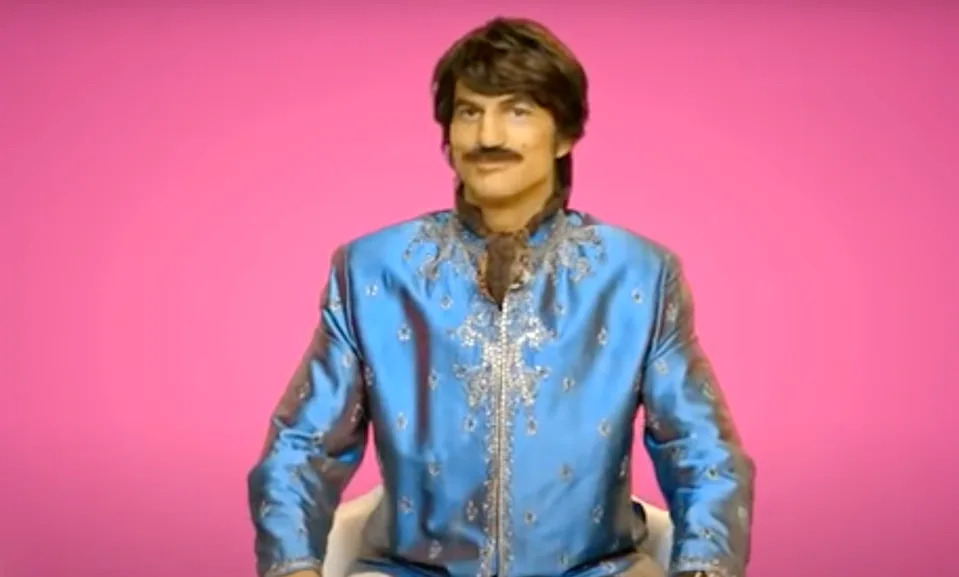 Person in ornate, patterned outfit with trimmed mustache seated against a pink background