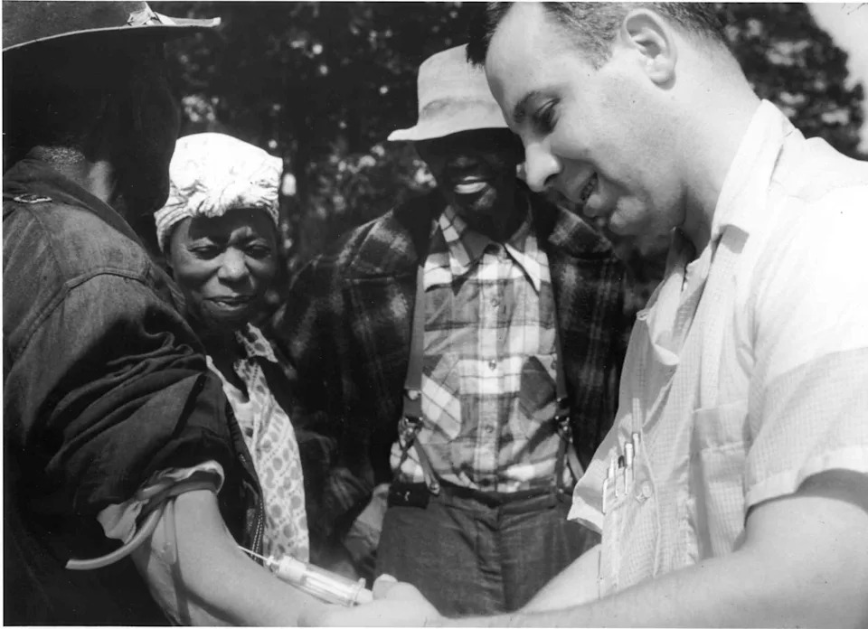 A healthcare worker administers a vaccine to a person's arm while others observe outdoors